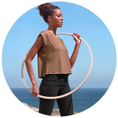 Frau in braunem Oberteil und schwarzen Hosen hält einen Hula-Hoop-Reifen am Strand mit Meer im Hintergrund