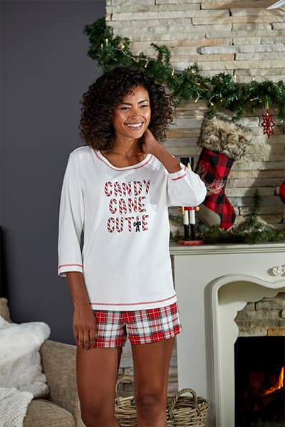 Candy Cane Cutie – Frau in weißem Shirt und karierten Shorts steht vor weihnachtlich dekoriertem Kamin