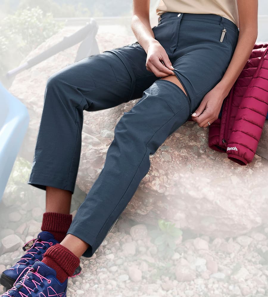 Frau sitzt auf Felsen und öffnet Reißverschluss an Wanderhose zum Abzippen des Hosenbeins