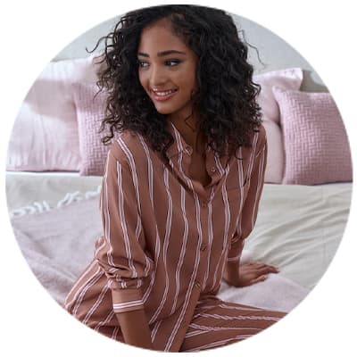 Frau mit lockigem Haar trägt gestreifte Pyjamas und sitzt auf einem Bett mit rosa Kissen