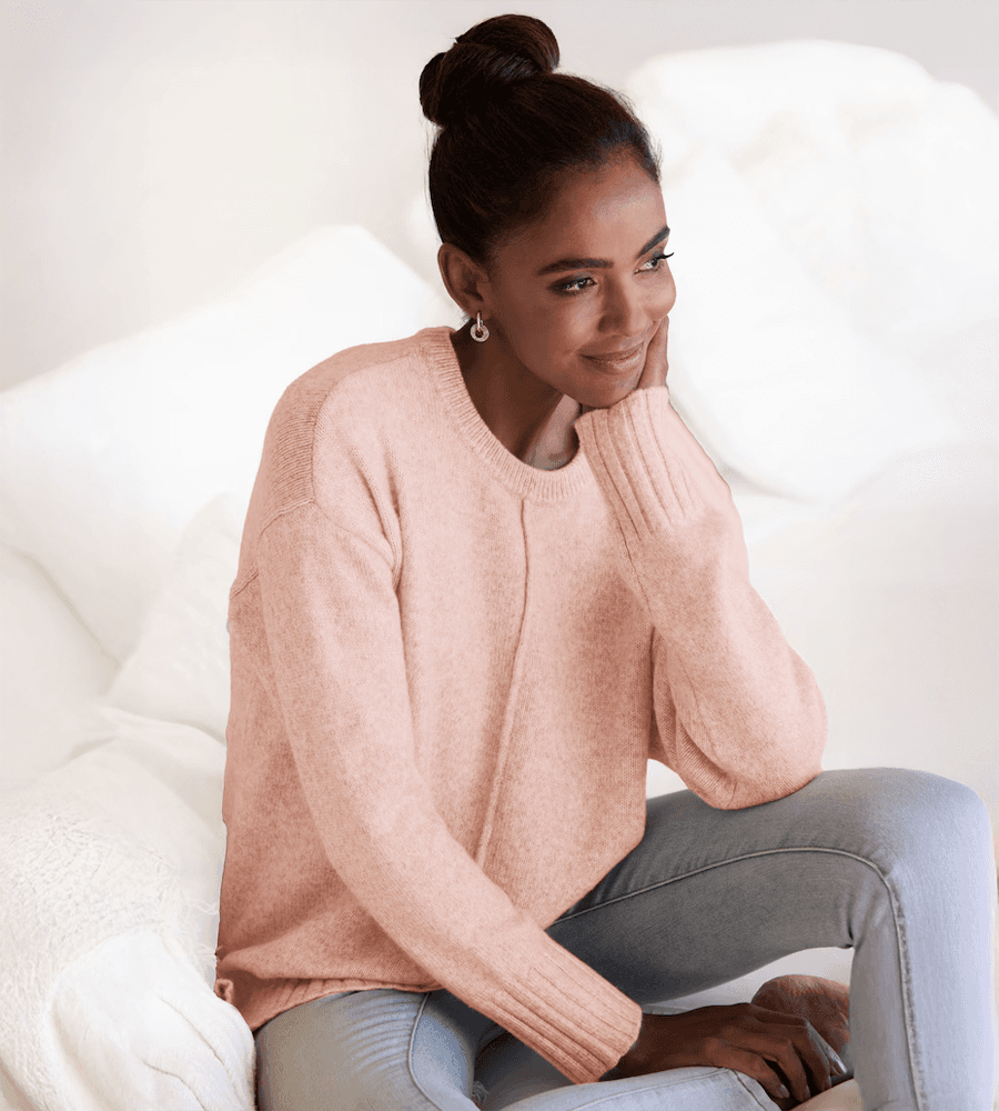 Frau trägt rosa Pullover und sitzt auf einem weißen Sofa, ihr Gesicht ist unscharf. Sie hat eine Hochsteckfrisur und silberne Ohrringe.