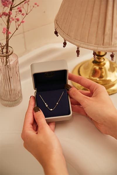 Hände halten eine Schmuckschatulle mit einer filigranen Halskette, daneben eine Tischlampe und eine Vase mit rosa Blumen