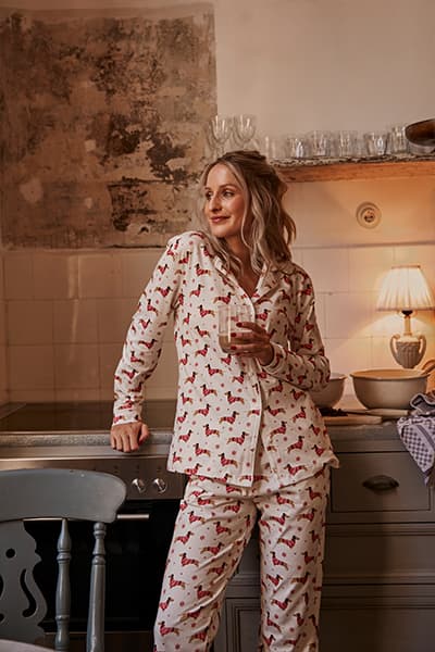 Frau in weißem Pyjama mit rotem Hundeprint, steht in der Küche und hält ein Glas, neben einem Stuhl und einer Lampe.