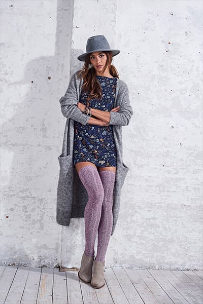 Frau in gemustertem Kleid, grauem Cardigan und lila Strümpfen, steht mit verschränkten Armen an einer Wand, trägt grauen Hut.