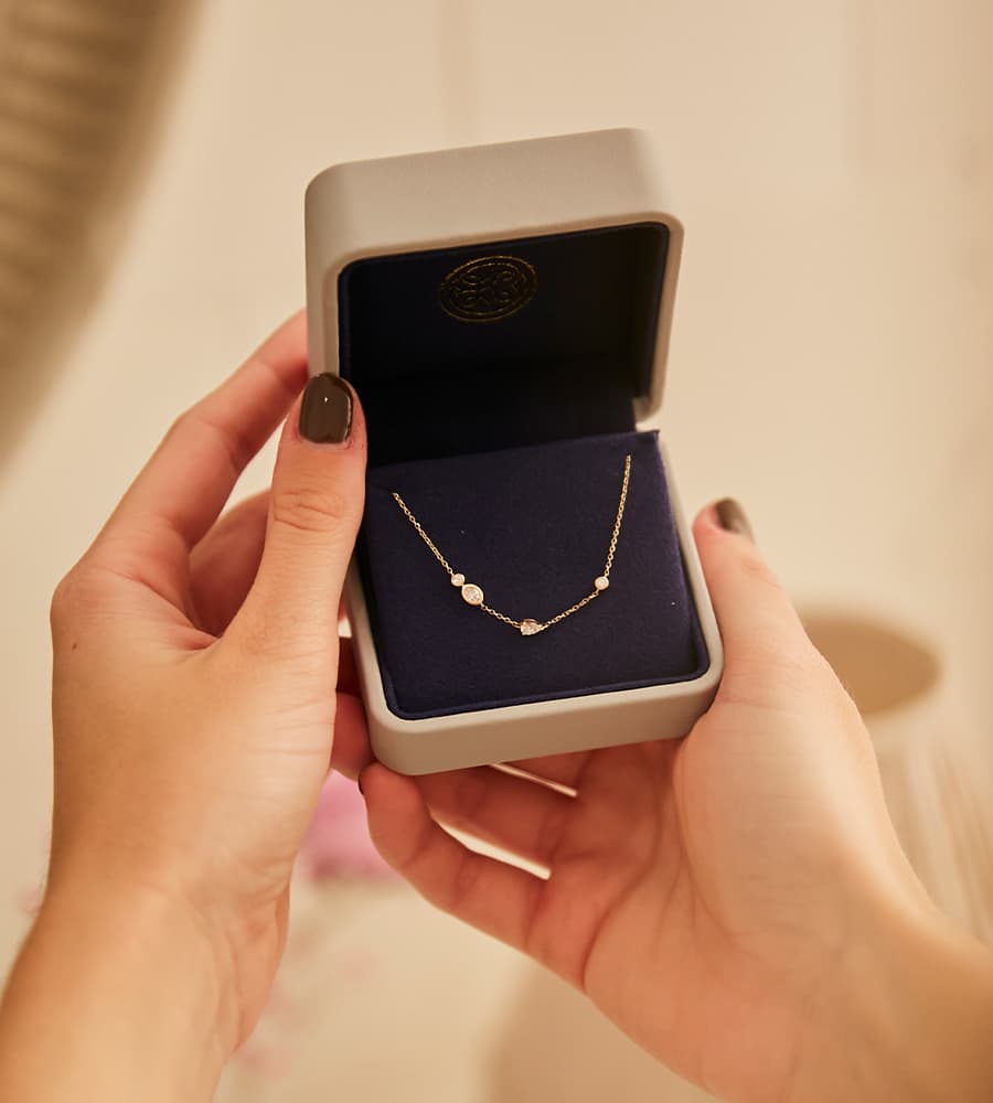 Hand hält eine Schmuckschatulle mit einer zarten goldenen Halskette mit drei kleinen Anhängern in verschiedenen Formen.