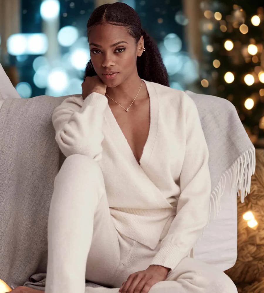 Frau in cremefarbenem Wickeloberteil und passender Hose sitzt auf einem Sofa vor einem festlich beleuchteten Hintergrund