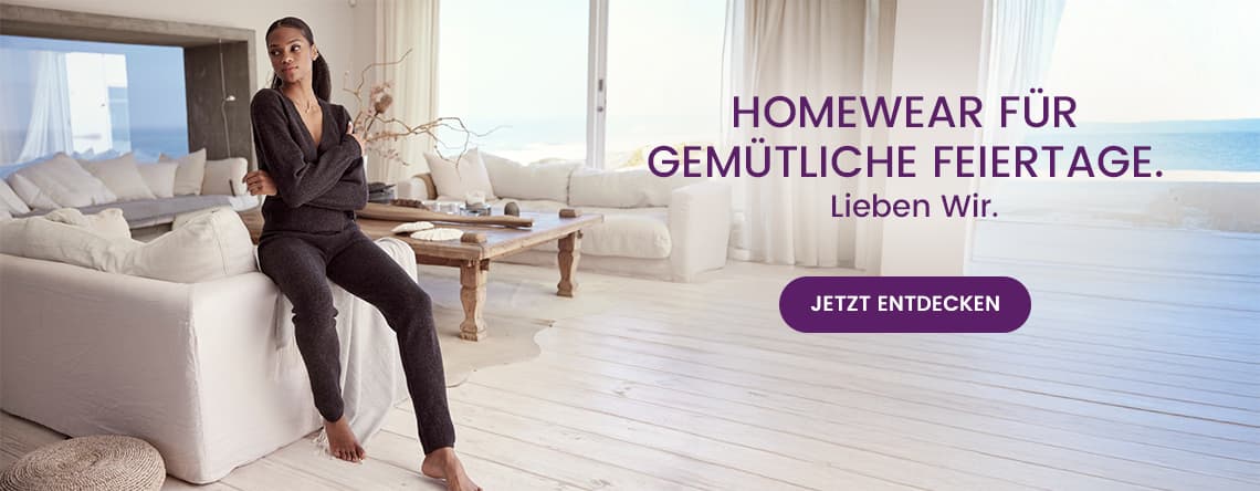 Homewear für gemütliche Feiertage. Lieben wir. Jetzt entdecken – Person in grauem Hausanzug sitzt entspannt auf weißem Sofa im hellen Wohnzimmer