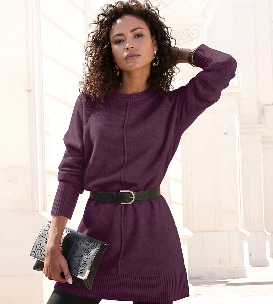Frau in lila Pullover mit schwarzem Gürtel und schwarzer Clutch, hält Arm hinter Kopf.