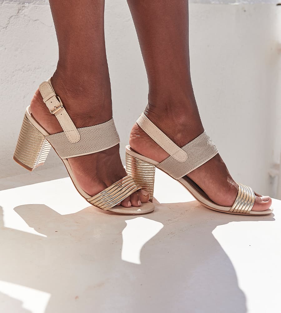 Füße in beigefarbenen Sandaletten mit Blockabsatz und Riemen, die im Sonnenlicht auf heller Fläche stehen