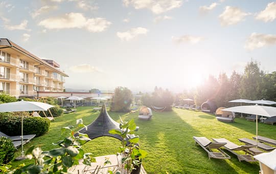 Hotelgarten mit Liegen, Sonnenschirmen und Loungekörben im Sonnenlicht vor mehrstöckigem Hotelgebäude