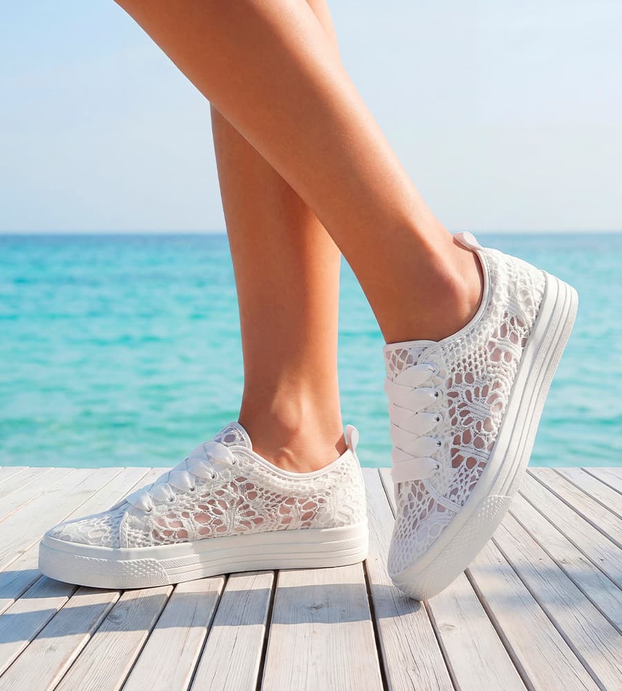 Füße in weißen Sneakern mit Lochmuster stehen auf Holzsteg vor blauem Meer