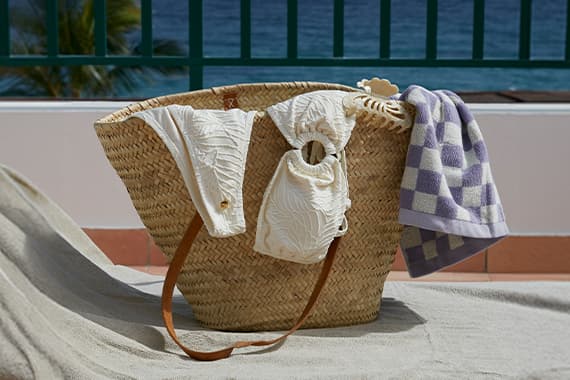 Große Strandtasche mit weißem Bikini und lila kariertem Handtuch auf Liege, im Hintergrund Meer und Balkonbrüstung