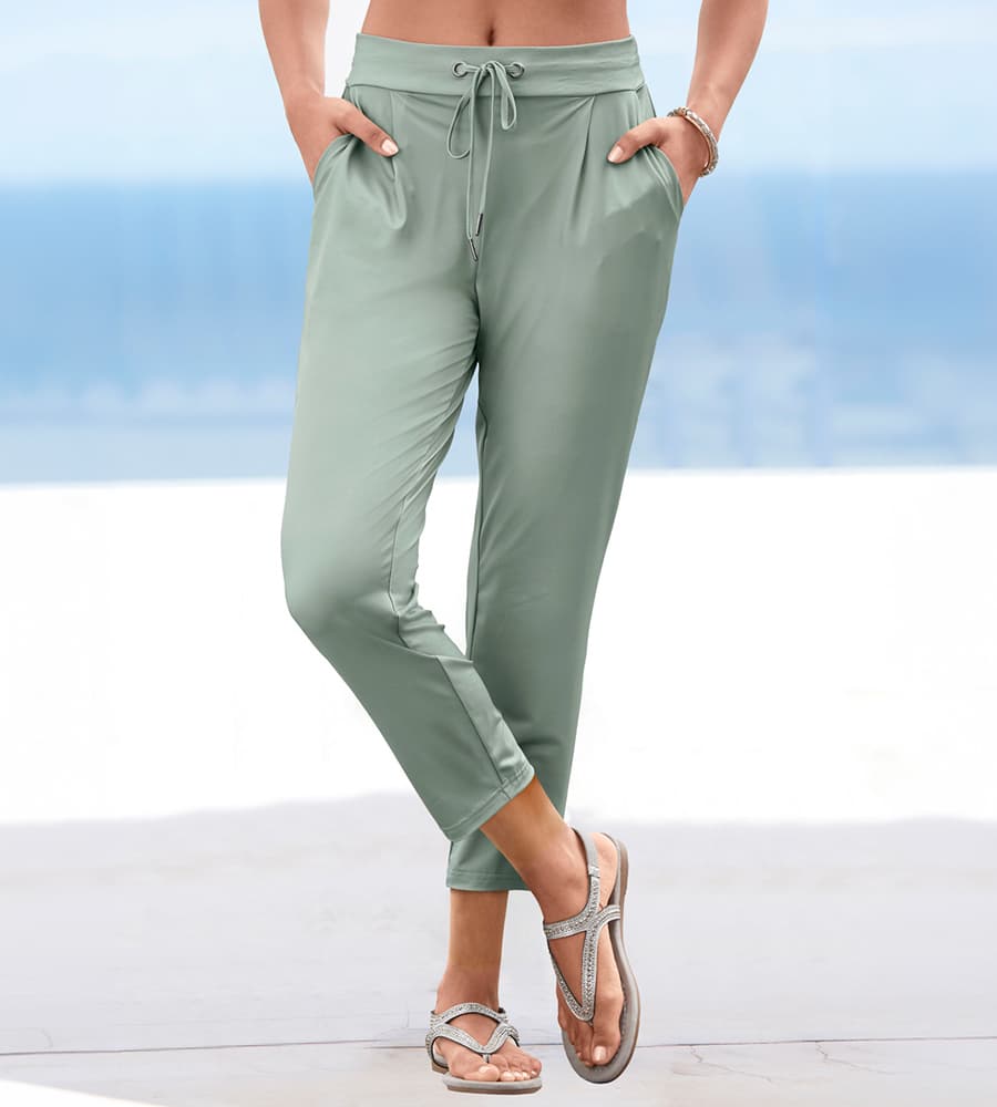 Frau am Strand in hellgrüner Jogpants mit Bindeband und Eingrifftaschen, dazu flache Sandalen