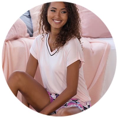 Frau im kurzen Schlafanzug mit rosa T-Shirt und gemusterter Shorts sitzt lächelnd auf einem Bett mit rosa Decke